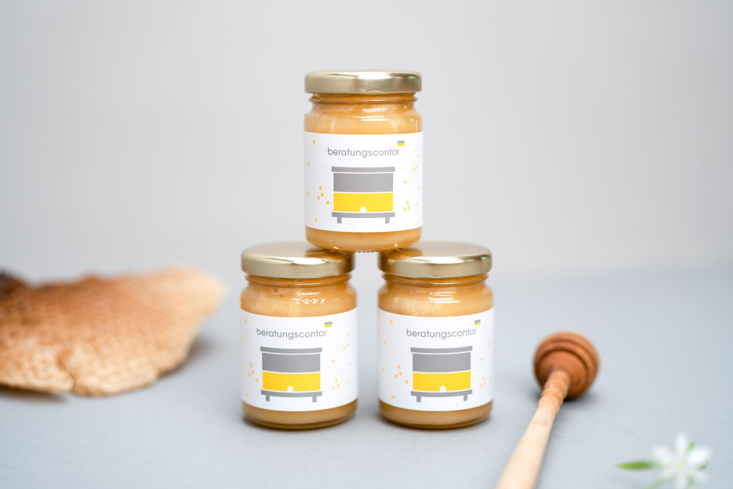 Drei gestapelte Honiggläser mit Etikett im minimaoistischen Beratungscontor-design, arrangiert auf einer grauen Fläche mit einem Honiglöffel und Bienenwaben im Hintergrund.