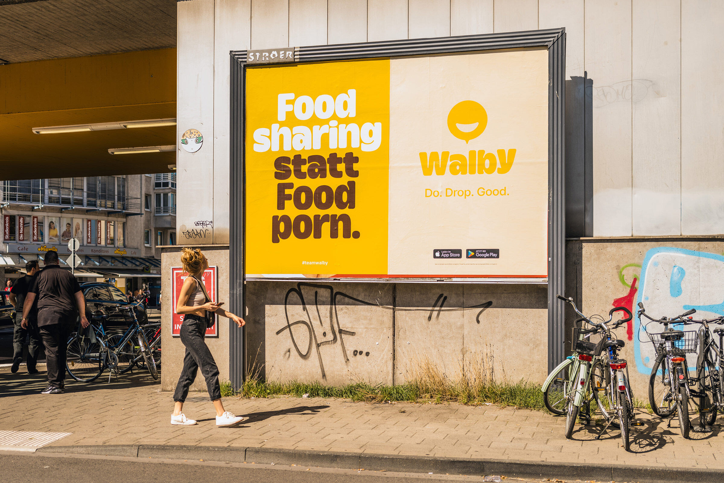 Großes Plakat der Walby-Launch-Kampagne unter einer Überführung in Köln. Links der Slogan ‘Food sharing statt Food porn.’ in weißer und brauner Schrift auf gelbem Hintergrund. Rechts das Walby-Logo mit einem lächelnden Kreis, der Schriftzug ‘Walby’ und der Claim ‘Do. Drop. Good.’ sowie die Logos von App Store und Google Play. Im Vordergrund geht eine Frau vorbei, und rechts sind Fahrräder abgestellt. Die Kampagne wirbt für nachhaltige und soziale Werte, passend zur Mission der Walby-App.