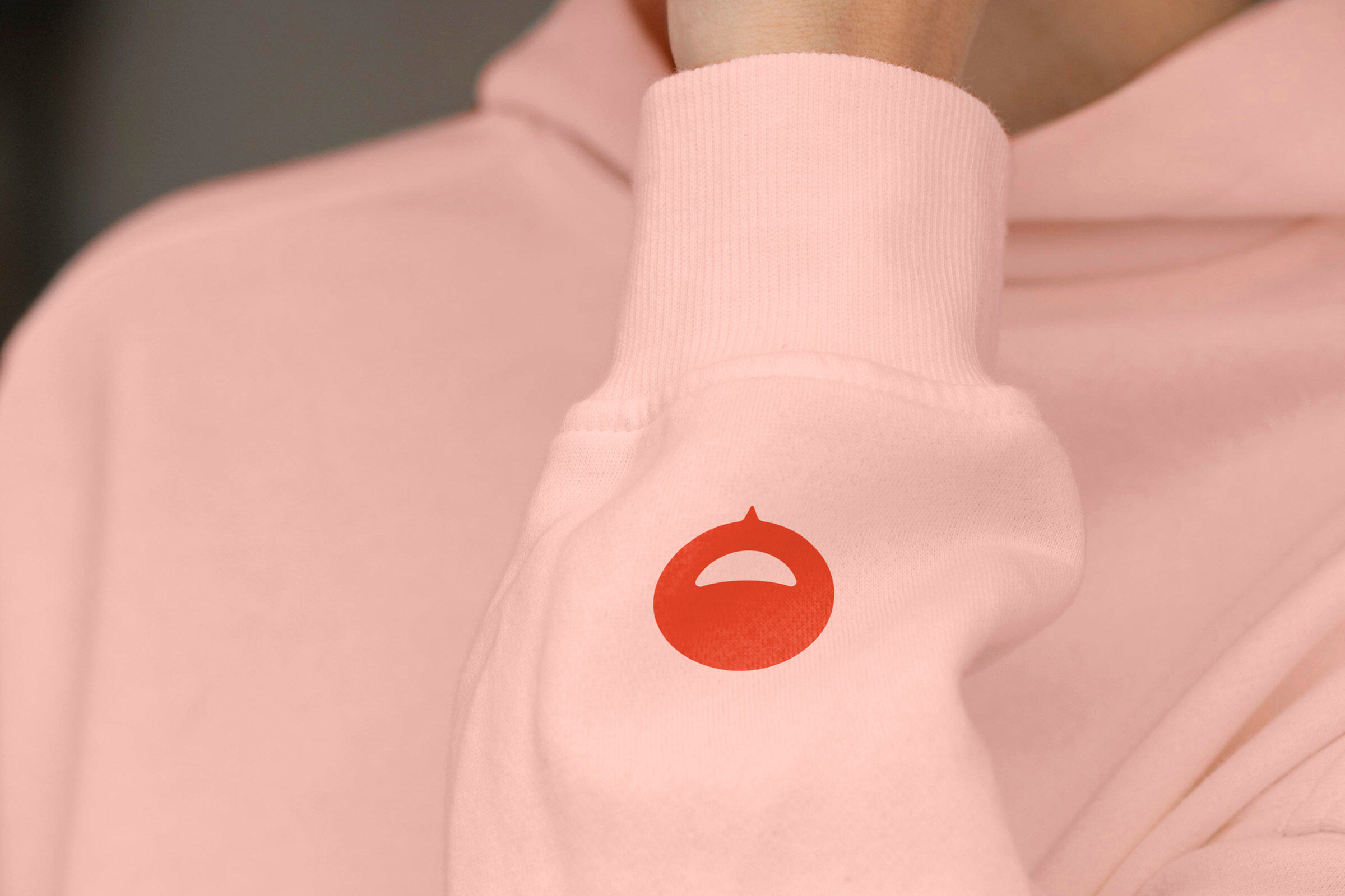 Detailaufnahme eines hellrosa Hoodies mit dem Walby-Logo auf dem Ärmel. Das rote Signet zeigt einen stilisierten Kreis mit einem lächelnden Mund, das die freundliche und positive Ausstrahlung der Marke Walby symbolisiert. Der Fokus liegt auf der hochwertigen und minimalistischen Gestaltung des Kleidungsstücks.