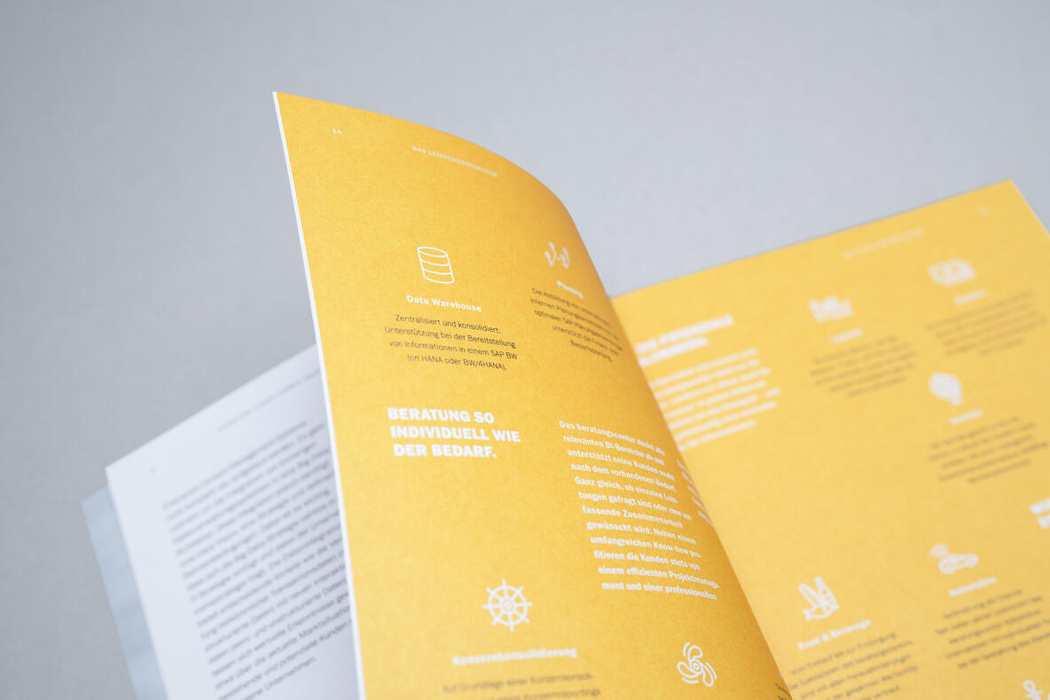 Geöffnete beratungscontor-Broschüre, gestaltet von hellofarm mit gelber Seite und weißen Icons sowie Textblöcken, die verschiedene Themen und Inhalte präsentieren, auf grauem Hintergrund fotografiert.