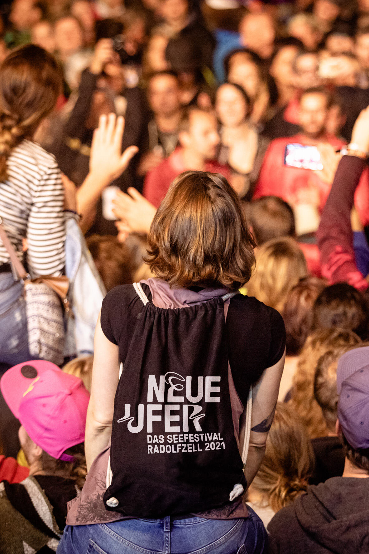 Frau mit einem schwarzen Stoffbeutel, bedruckt mit der „Neue Ufer – Das Seefestival Radolfzell 2021“, inmitten einer Menschenmenge bei einem Festival.