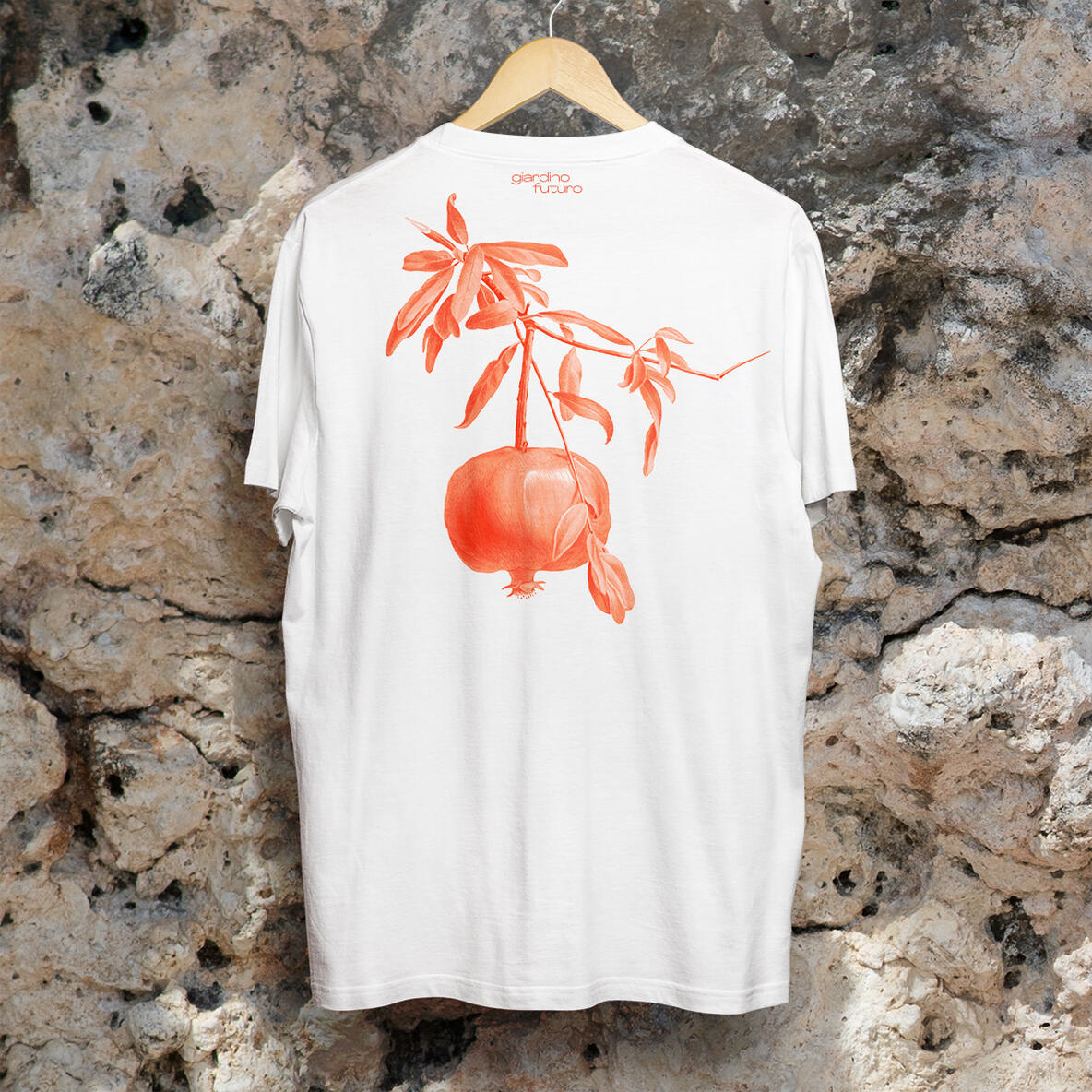 Rückseite eines weißen T-Shirts mit einer Illustration eines Granatapfelzweigs in leuchtendem Rot-Orange, aufgehängt vor einer rustikalen Steinwand.