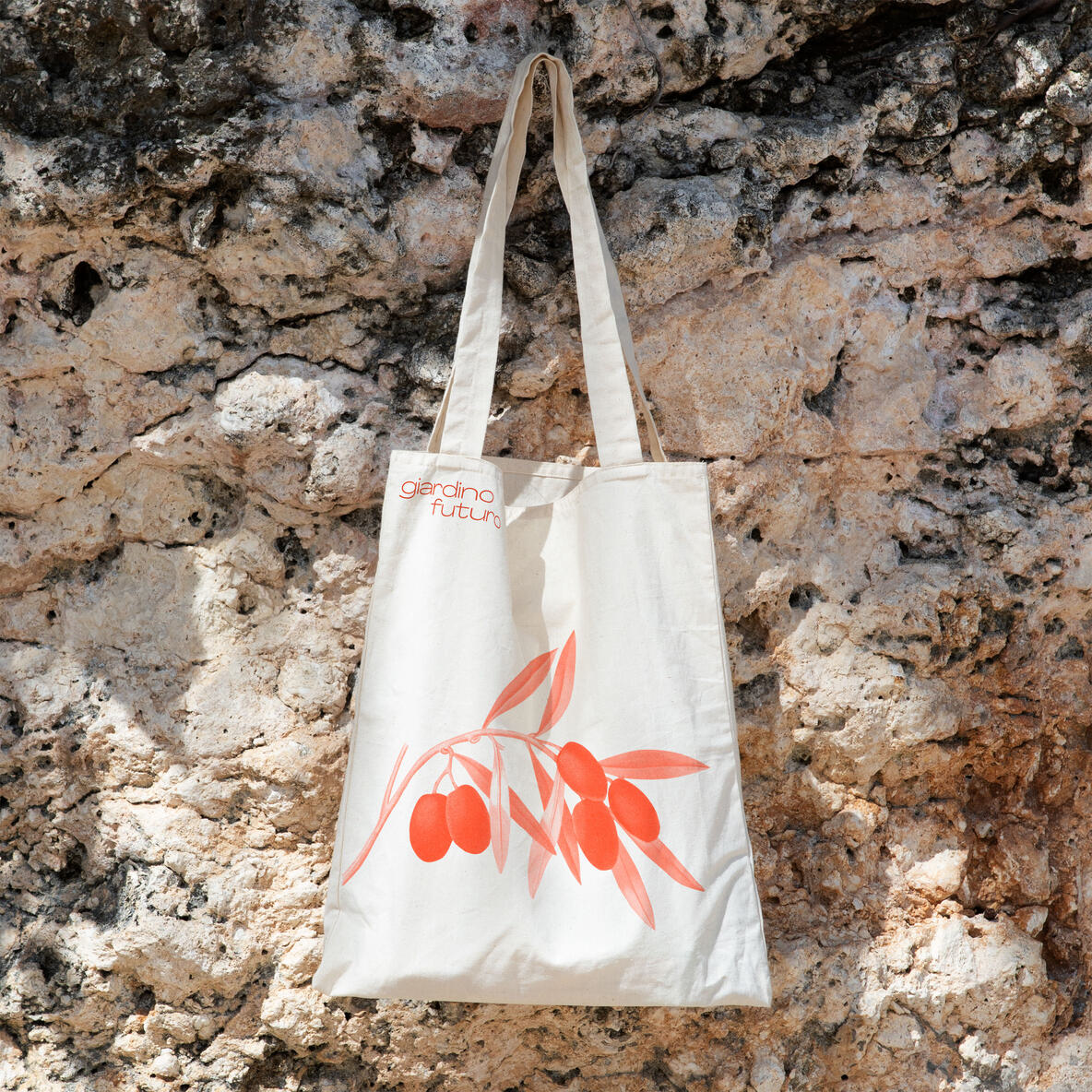 Leichte Stofftasche mit Olivenzweig-Illustration und dem Schriftzug ‘Giardino Futuro’, aufgehängt vor einer rustikalen Steinwand.