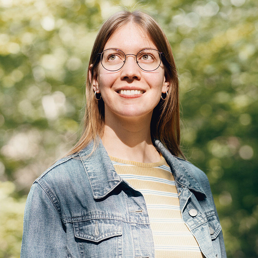 Porträt von Annika Bitzer mit Brille, die lächelnd in die Ferne schaut, umgeben von einer natürlichen, grünen Hintergrundszenerie.