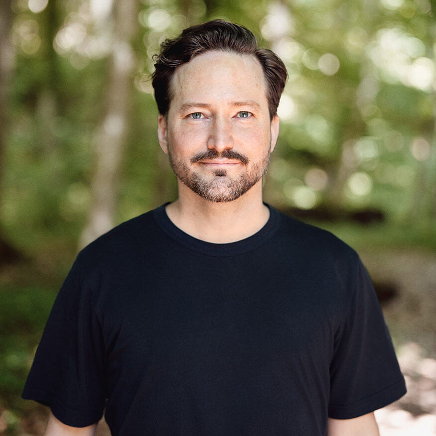 Porträt von Björn Küenzlen mit kurzen Haaren, Bart und einem schwarzen T-Shirt, der in einem sonnendurchfluteten Wald steht.