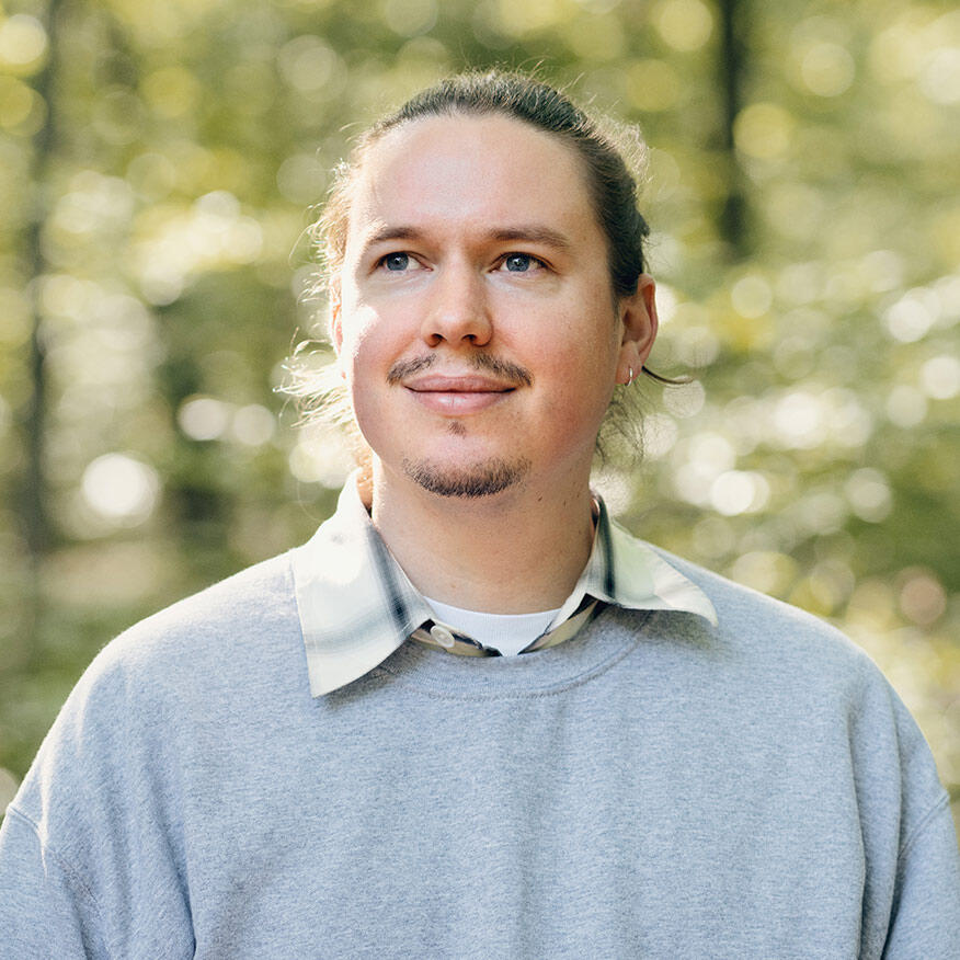 Porträt von Robin Christopher Körner, der lächelnd in die Ferne schaut, umgeben von einer natürlichen, grünen Hintergrundszenerie.