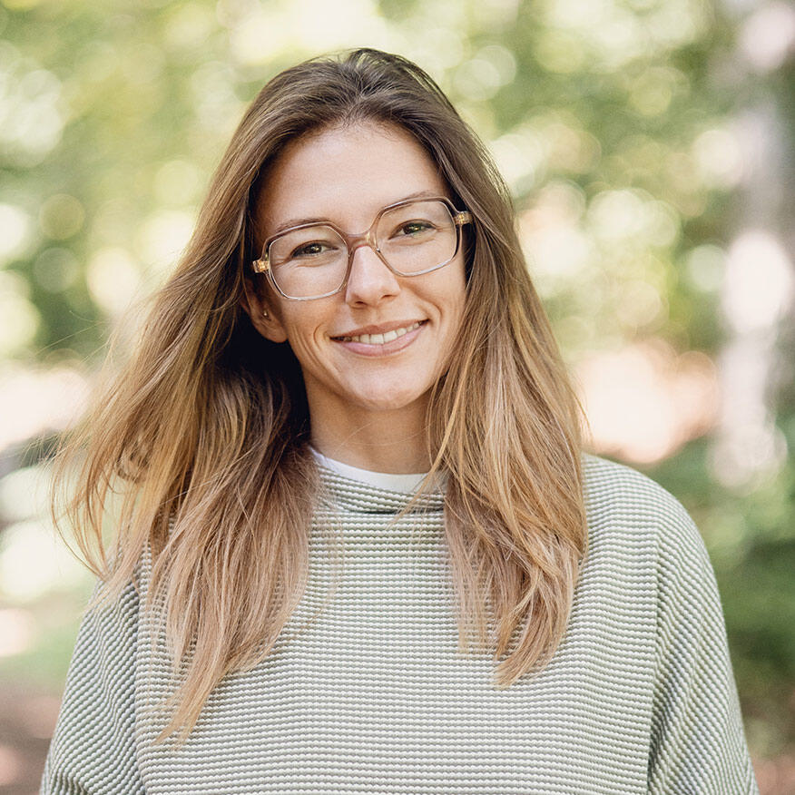 Porträt von Tina Zeltwanger, die lächelt und eine Brille, langes offenes Haar und einen gestreiften Pullover trägt, vor einem natürlichen, grünen Hintergrund.