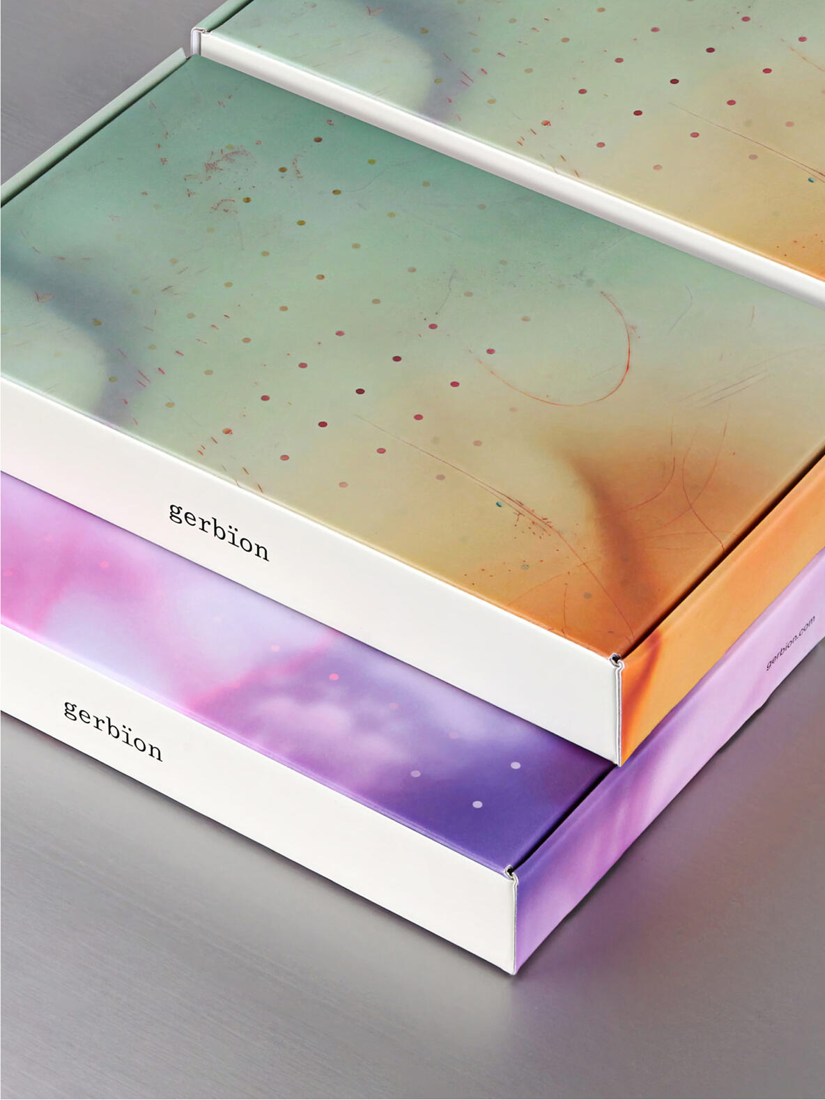 Zwei flache Produktverpackungen von gerbion liegen übereinander auf einer grauen Oberfläche. Die Boxen zeigen die charakteristischen, organischen Farbverläufe in Grün-Orange und Violett-Rosa, kombiniert mit einem dezenten Punktraster. Der Markenname „gerbion“ ist jeweils minimalistisch auf dem weißen Seitenrand platziert.