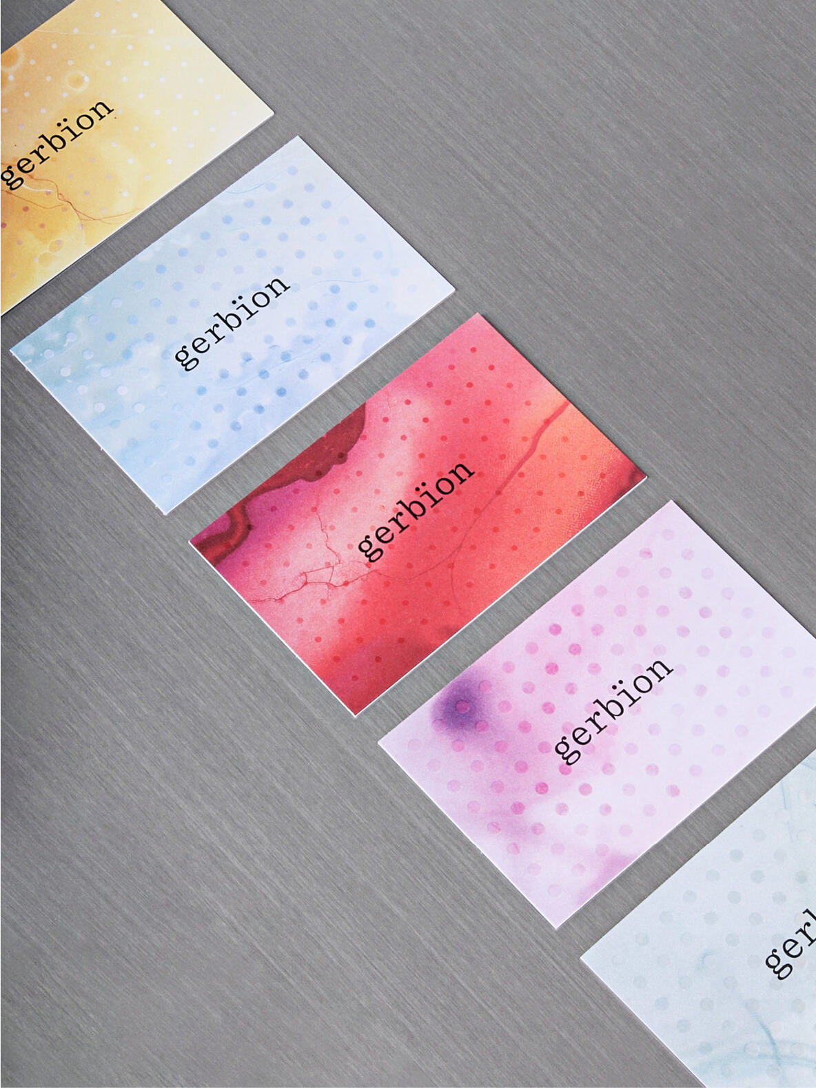 Sechs gerbion-Visitenkarten in unterschiedlichen Farbvarianten – Gelb, Hellblau, Rot, Rosa, Lila und Türkis – liegen fächerförmig auf einer grauen Metalloberfläche. Die Karten zeigen das gerbion-Logo in Monospace-Typografie, kombiniert mit einem Punktmuster und organischen Farbverläufen, inspiriert von mikrobiologischen Strukturen.